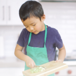 A young boy in a blue t-shirt and green aprons scrapes the vegetables he chopped off a cutting board and into a bowl.